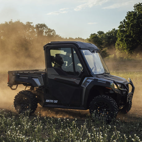 A black Arctic Cat side-by-side UTV with a canopy is driving through a dusty field, kicking up a cloud of dirt. Trees and a blue sky are visible in the background.