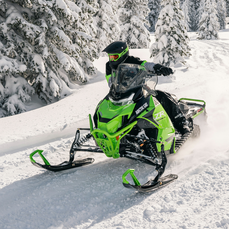 A person riding a green and black Arctic Cat snowmobile through deep snow, with snow-covered pine trees in the background.